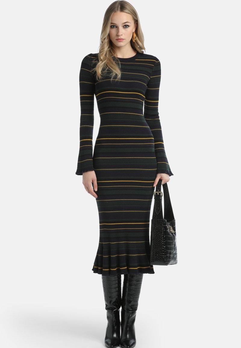 Juoda ir geltona dryžuota raštuota trikotažo suknelė su varpelio formos rankovėmis ir išplatintu apvadu, derinama su juodais patentiniais batais iki kelių ir krokodilo tekstūros krepšiu.