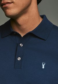 Tummansininen polo-paita, jossa on teksturoitu kaulus, kolme nappia etuosassa ja vaaleansininen brodeerattu peuran logo vasemmalla rinnalla.