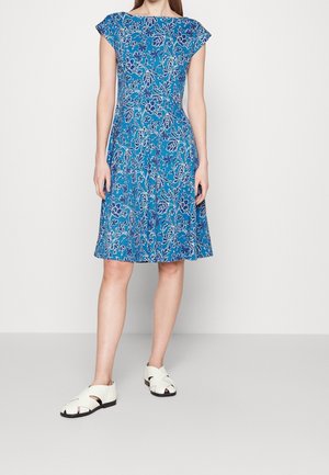 Vrouw in een knie-lange blauwe jurk met wit bloemmotief en witte open-toe sandalen, staand tegen een effen lichte achtergrond.