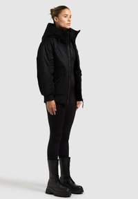 Giacca a vento nera con colletto alto, chiusura a zip e tasche laterali. Indossata con leggings neri e stivali da combattimento con lacci. Texture liscia.