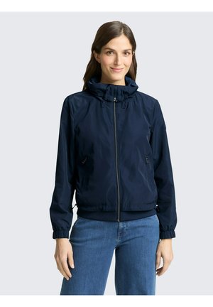 Frau trägt eine marineblaue Jacke mit Reißverschluss, hohem Kragen und elastischen Bündchen, kombiniert mit blauen Jeans, stehend vor einem schlichten Hintergrund.