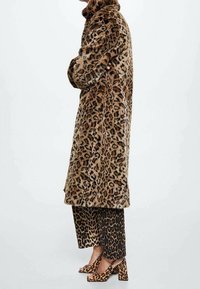 Cappotto in pelliccia sintetica con stampa leopardata, di lunghezza media, con colletto alto; abbinato a pantaloni coordinati con stampa leopardata e tacchi grossi. Tessuto texturizzato.