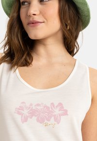 Top bianco senza maniche con stampa floreale rosa e scritta dorata 'Roxy'. Tessuto morbido con scollo rotondo e ampie aperture per le braccia. Cappello verde sullo sfondo.