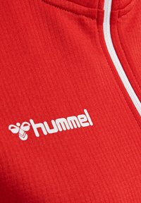 Giacca sportiva rossa con texture a coste, dotata di zip bianca e un logo bianco con la scritta "hummel" sulla parte frontale.