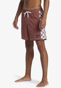 Men's swim trunks in burnt orange with a checkerboard pattern on one side. Features a drawstring waist and logo patch. Fabric appears lightweight.