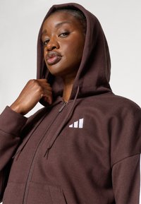 Smešani smeđi zip-up dukserica s teksturiranom tkaninom, koja ima bijeli logo na lijevoj strani prsnog područja i kapuljaču s vezicom.