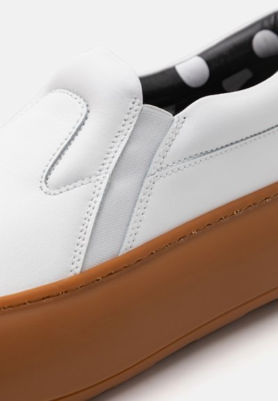 White leather slip-on shoe with elastic side panels, brown rubber sole, and a textured finish. Interior features black with white polka dots.