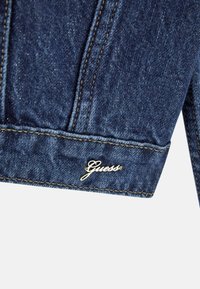 Mörk denimjacka med en gyllene "Guess"-logotyp vid fållen, med synlig sömnad och en texturerad tygfinish.