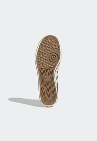 Podešev tenisky z hnědé gumy s texturovaným povrchem. Obsahuje kruhové a kosočtverečné vzory a bílý boční pruh. Viditelné výrazné logo.