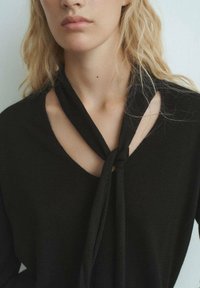 Top nero lavorato a maglia con scollatura a V e dettagli di una sciarpa annodata al colletto, con una texture liscia e scollatura aperta.