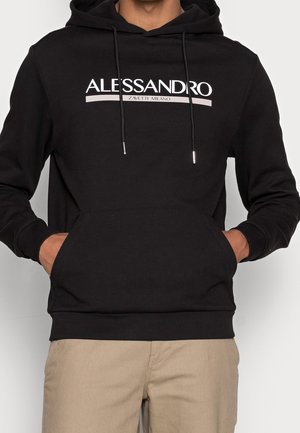 Personne portant un sweat à capuche noir avec le texte "ALESSANDRO ZAVETTI MILANO", les mains dans la poche avant, associé à un pantalon beige.