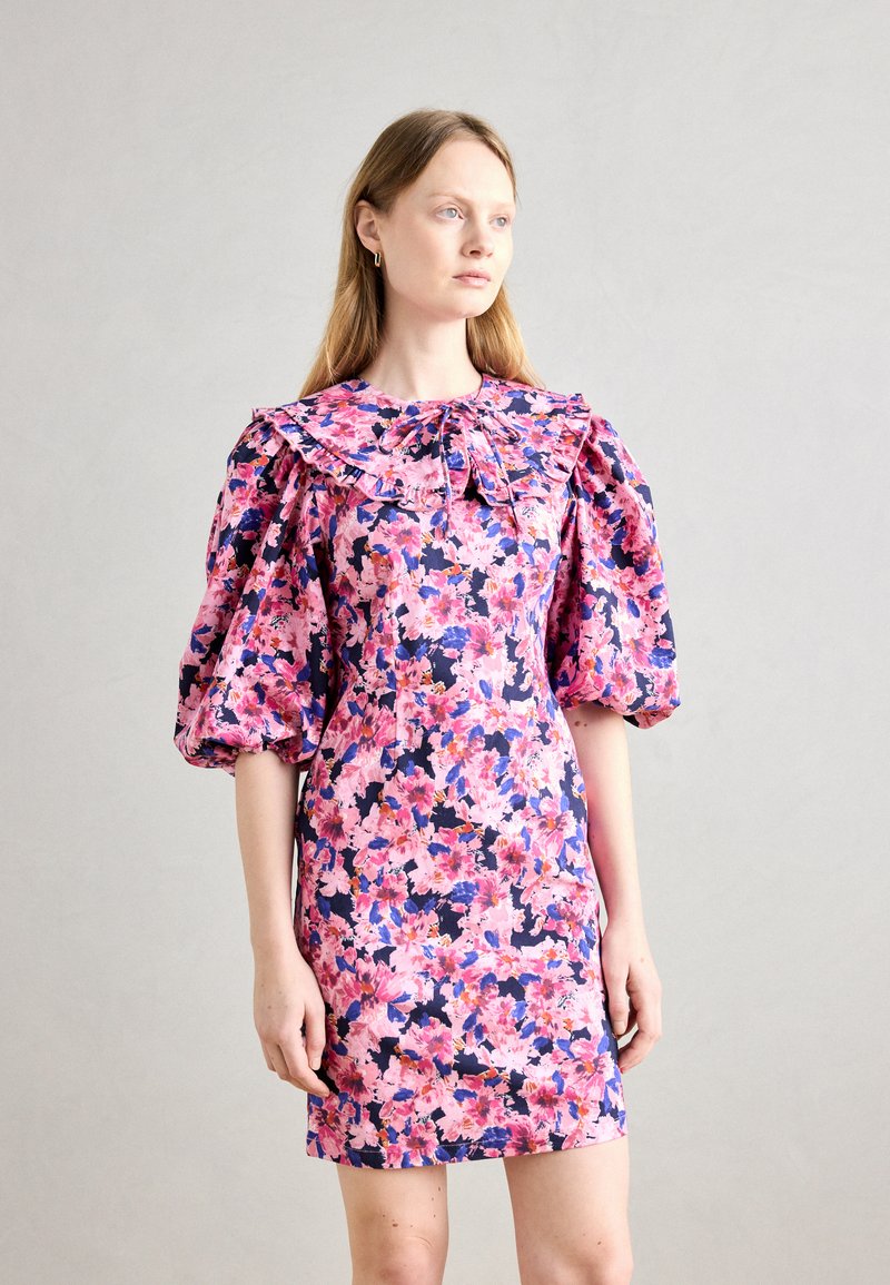 Robe florale avec des manches bouffantes, un col volanté et une silhouette cintrée. Les couleurs incluent des motifs floraux roses, violets et bleus sur un fond marine.