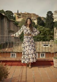 Maxikleid in Creme mit schwarzen Blättern, langen Ärmeln und tailliertem Schnitt. Gestylt mit einem braunen Gürtel und weichen braunen Schuhen.