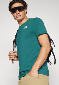 Turkusowa koszulka z krótkim rękawem z logo The North Face, noszona z khaki spodniami i czarnym plecakiem. Model w ciemnych okularach przeciwsłonecznych.
