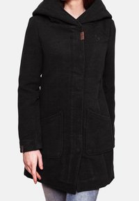 Femme portant un manteau noir à capuche avec poches sur le devant, une fermeture à l'avant et une broderie de logo discrète sur la poitrine.