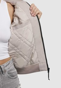 Heled beež bomber-jakk, tikandiga vooder, soonikkoes, küljetaskud, lukuga sulgemine ning sile tekstuur. Sisemise voodri pind on läikiv ja pehme.