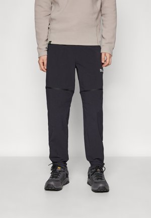 Person wearing black zip-off hiking pants and gray hiking shoes standing against a plain background.