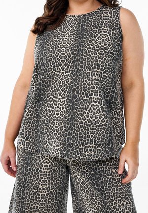 Femme portant un haut sans manches et un pantalon large à motif léopard noir et beige, debout devant un fond blanc.