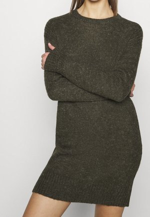 Femme portant une robe pull tricotée moulante à manches longues de couleur vert olive foncé avec un col rond, debout les bras croisés.