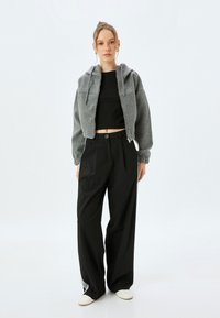 Giacca in pile grigia, tagliata, con cappuccio, abbinata a pantaloni neri a vita alta e gamba larga e scarpe bianche; design minimalista, texture morbida.