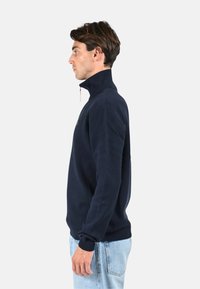 Sweatshirt zippé bleu marine avec col haut, poignets côtelés et texture douce. Présente un logo discret sur la gauche de la poitrine et une coupe décontractée.