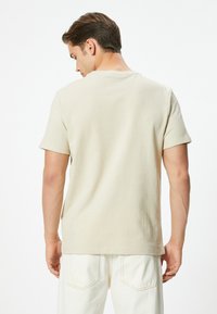 Beige kortærmet T-shirt lavet af struktureret stof, med rund hals og enkelt design, set bagfra med hvide bukser.