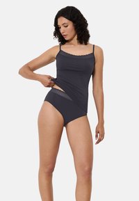 Kobieta ubrana w ciemnoszary top na ramiączkach z koronkowym wykończeniem oraz dopasowane wysokie koronkowe majtki, delikatnie podnosząca top, aby pokazać szczegóły pasa.