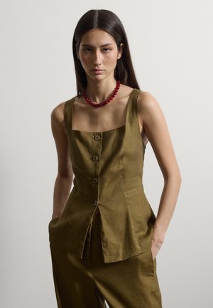 Donna con capelli lunghi e scuri, in piedi con le mani nelle tasche, indossa un top senza maniche verde oliva con bottoni e pantaloni abbinati, e una collana rossa di perline.