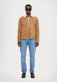 Brun ruskindsjakke med en pelskrave, frontlynlås, to synlige lommer, parret med lyseblå jeans og sorte sko.