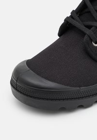 Zapatilla deportiva negra con una parte superior de tela texturizada, puntera de caucho y suela de caucho con un patrón. Presenta cordones y ojales negros.