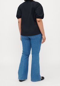 Schwarzes Oberteil mit Puffärmeln und rundem Ausschnitt, kombiniert mit blauen Schlagjeans. Das Outfit zeichnet sich durch eine glatte Textur und eine figurbetonte Silhouette aus.