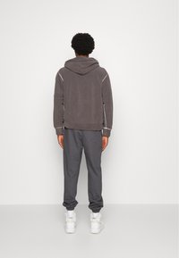 Felpa grigia con zip e cappuccio, abbinata a pantaloni jogger grigi. Presenta polsini elasticizzati e una vestibilità rilassata, con cuciture bianche a contrasto. Sneakers bianche.