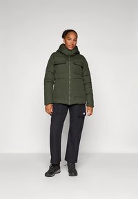 Žena stoji u tamnozelenoj puffer jakni sa kapuljačom, crnim cargo hlačama i crnim planinarskim čizmama na jednoličnoj svjetloj pozadini.