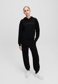 Zwarte cropped hoodie met geborduurd logo, gecombineerd met bijpassende wijde sweatpants en witte platform sneakers. Glad materiaal, minimalistisch ontwerp.