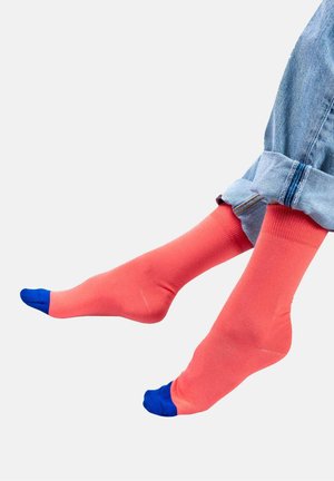 Koralowe skarpetki z niebieską osłoną na palce, o gładkiej fakturze. Noszone z podwiniętymi niebieskimi dżinsami, ukazując intensywny kontrast kolorów.