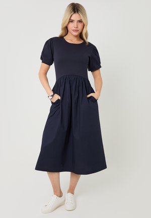 Vrouw staand met handen in de zakken, draagt een marineblauwe midi-jurk met pofmouwen en witte sneakers, tegen een effen achtergrond.