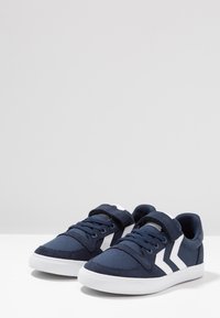 Navy canvas sneakers with white accents, featuring lace-up and Velcro closure, flat white rubber soles, and a rounded toe design.