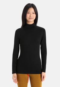 Sort turtleneck-top lavet af glat stof med lange ærmer, tætsiddende design og høj krave. Sammen med sennepsfarvede bukser med en rød snor.
