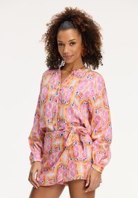 Kleurrijke, gestreepte jurk met een wikkelriem, gemaakt van lichte stof. Heeft lange mouwen en een kraag, voornamelijk roze met gele accenten.