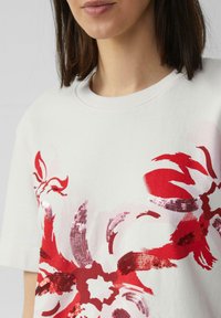 Weißes Baumwoll-T-Shirt mit kurzen Ärmeln, das ein großes rotes florales Paillettenmuster und einen gerippten Halsausschnitt aufweist. Weicher Stoff mit lockerer Passform.