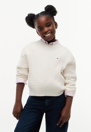 Junges Mädchen mit zwei Puff-Frisuren, trägt einen weißen Zopfpullover über einem rosa Hemd und dunkelblauen Jeans, lächelnd mit leichtem Kopfneigen.