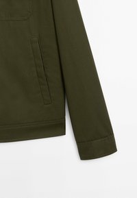 Manche et partie inférieure d'une veste vert olive avec une poche verticale à l'avant et des coutures visibles sur fond blanc.