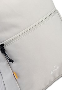 Lichtgrijze rugzak van gestructureerde stof, met een ritsluiting, gemarkeerde ritsen en een subtiele logo aan de zijkant.