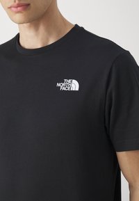 Detailní záběr na osobu v černém tričku s bílým logem "The North Face" na levé straně hrudníku.