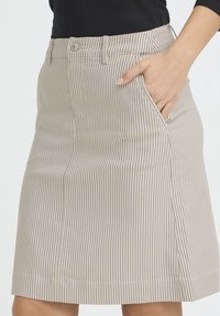 Nærbillede af en person iført et beige nederdel med tynde lodrette striber og en hånd placeret i forlommen.