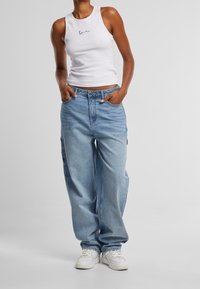 Bílé žebrované tílko s malým černým logem, kombinované s modrými volnějšími džíny a bílými teniskami, které představují ležérní, uvolněný styl.