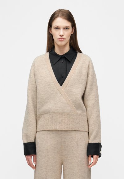 Femme portant un pull beige portefeuille par-dessus une chemise noire à col et un pantalon beige assorti, debout devant un fond blanc uni.