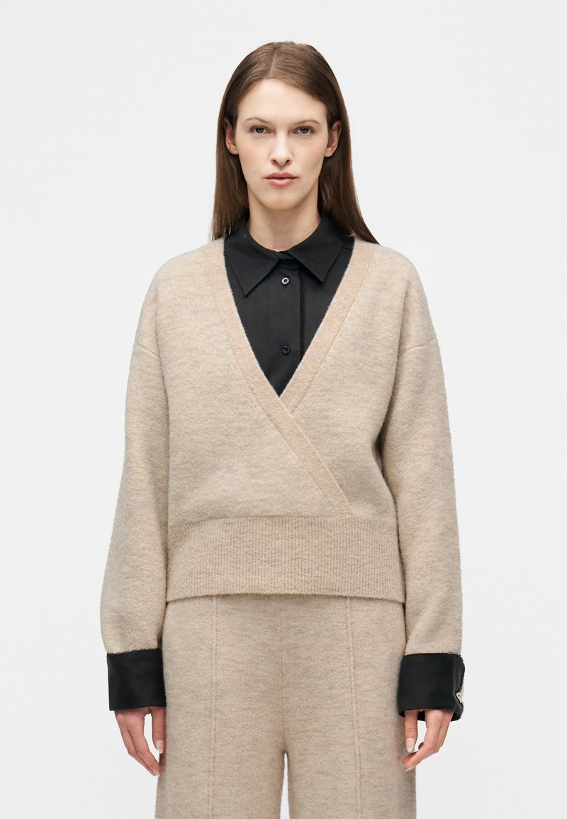 Γυναίκα που φοράει μπεζ πουλόβερ τ ύπου wrap πάνω από μαύρο πουκάμισο με γιακά και μπεζ παντελόνι που ταιριάζει, στέκεται μπροστά σε απλό λευκό φόντο.