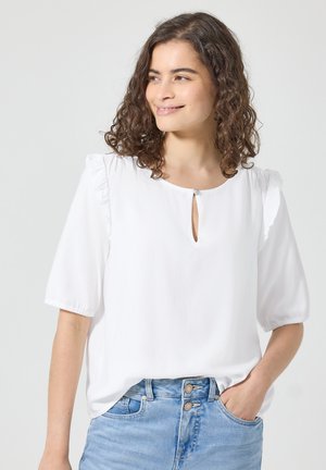 Kvinde med krøllet hår, der smiler, iført en hvid bluse med korte ærmer og en lille åbning foran samt lyseblå jeans, hånd i lommen.