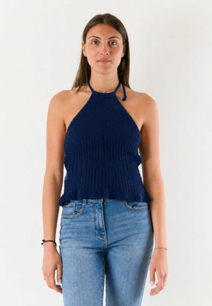 Donna con capelli lunghi e scuri che indossa un top halter blu navy lavorato a maglia e jeans azzurri, in piedi su uno sfondo bianco.